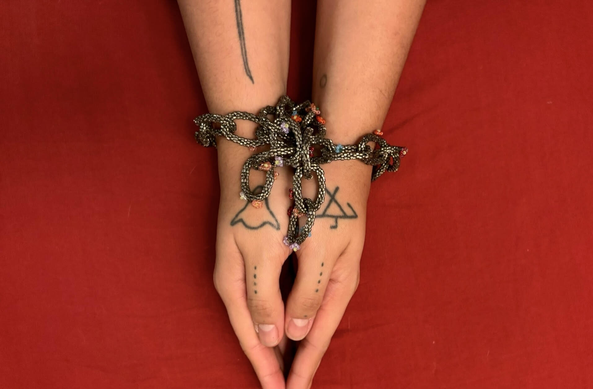 Woman's hands with Indigenous tatoos, wrists bound together by a beaded chain.